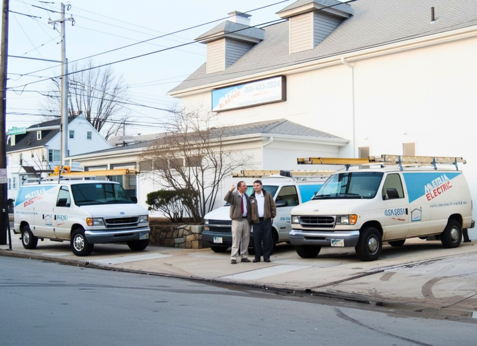 Two white service vans parked outside a house, with three people standing between them on a snowy street.