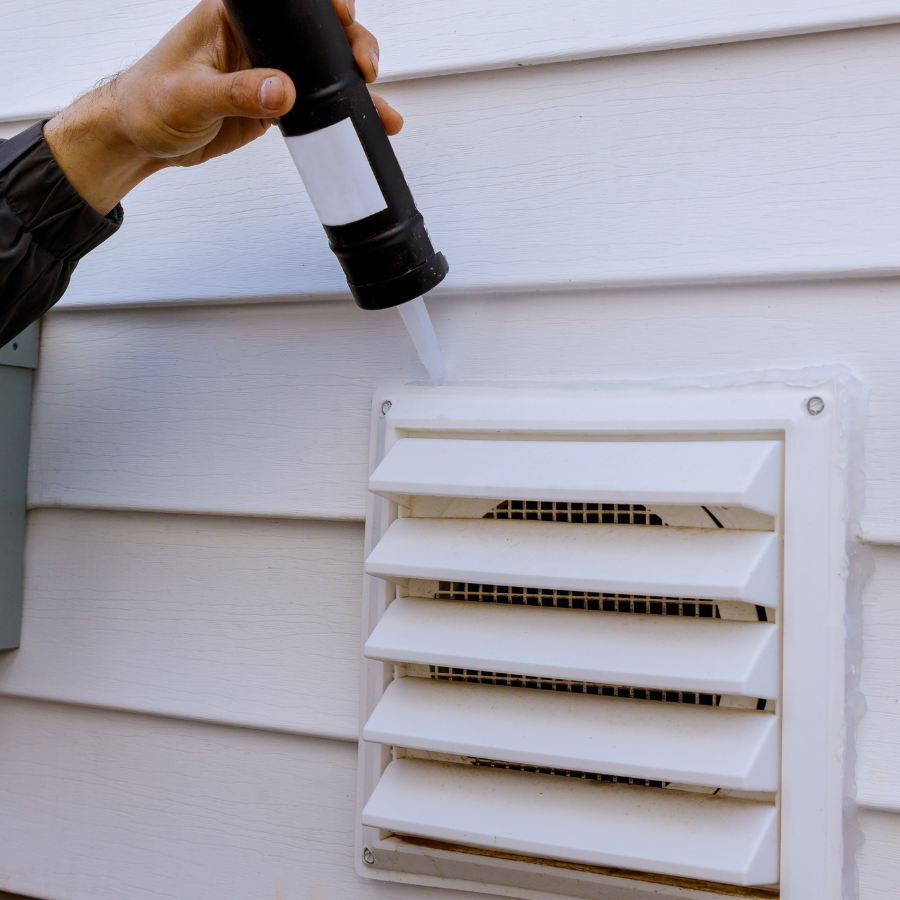 Hand holding a flashlight near a white exterior wall vent with louvered slats