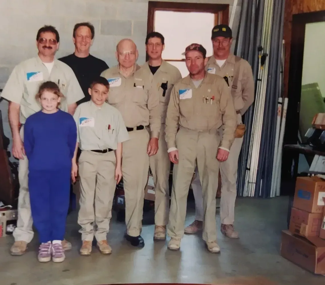 Group standing indoors, with two children in front and several adults in tan uniforms behind them
