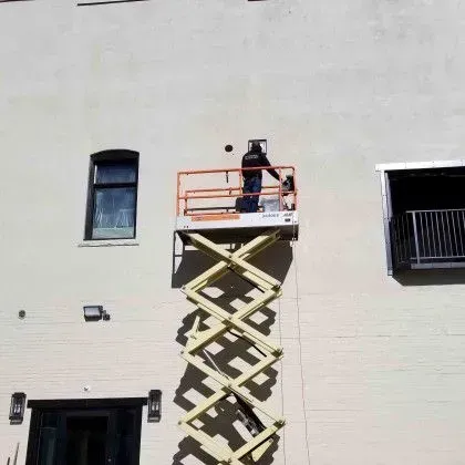 Worker on an elevated scissor lift outside a beige building, near a window and balcony.