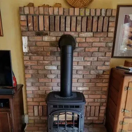 Black wood stove with tall pipe in front of a brick fireplace wall in a living room.