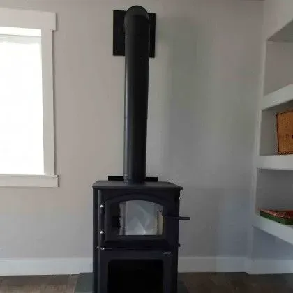 Black wood stove with a tall chimney in a light gray room.