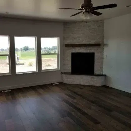 Empty living room with dark wood floors, large windows, and a white brick fireplace with a ceiling fan.