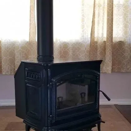Black cast-iron wood stove with a glass door and chimney pipe in front of curtained windows