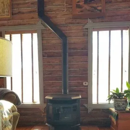 Rustic cabin interior with wood stove, tall windows, and a potted plant on a side table.