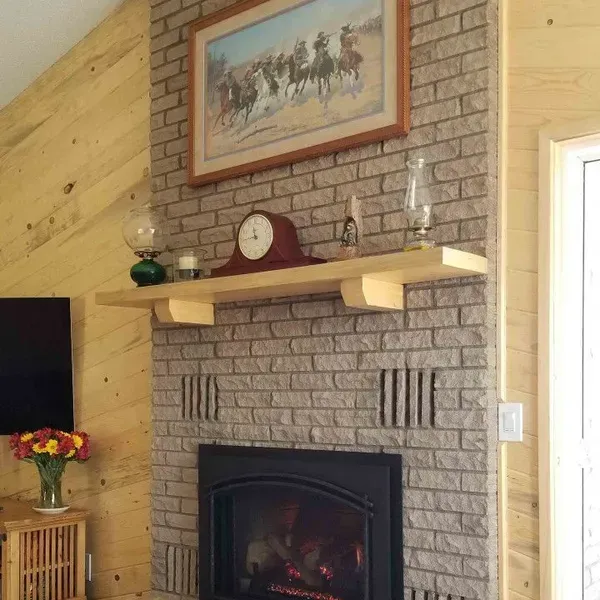 Living room fireplace with stone surround, wooden mantel, framed painting, and clock against a brick wall.