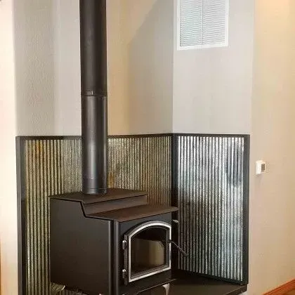 Black wood-burning stove with a chimney pipe in a room corner, beside a curved black metal heat shield.