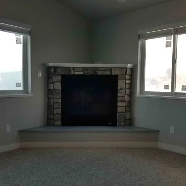 Empty living room with stone fireplace, two windows, and carpeted floor.