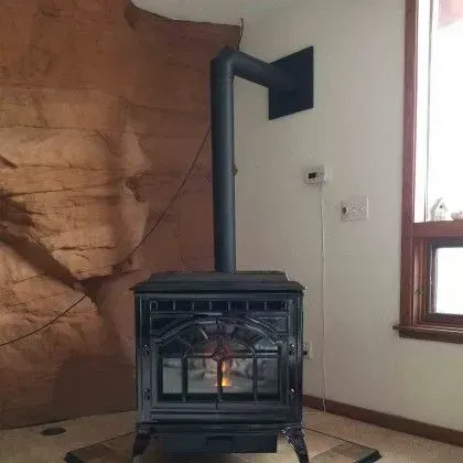 Black wood stove with glowing fire in a room beside a window and stone wall