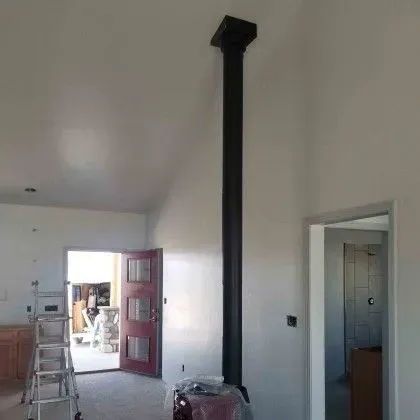 Bright unfinished room with a tall black chimney pipe, open doorway, ladder, and kitchen area in the background