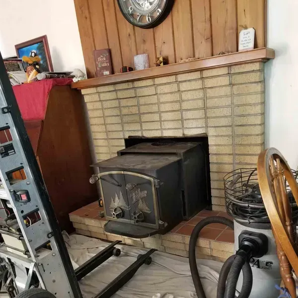 Wood-paneled living room with a brick fireplace, black wood stove, and vacuum cleaner in the foreground
