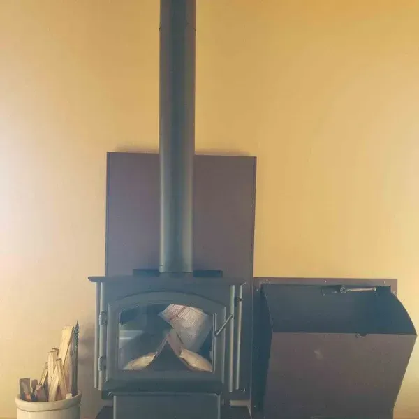 Black wood stove with chimney against a beige wall, with firewood stored in a bin beside it