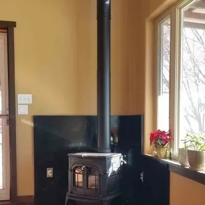 Wood-burning stove with black chimney pipe in a corner, beside a window and potted plants.