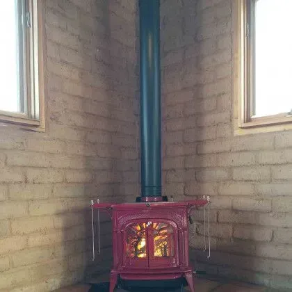 Red wood-burning stove with a glowing fire in a corner between two windows, with a blue flue pipe above.