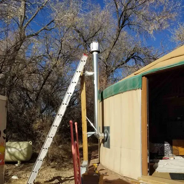 Ladder leaning against a metal vent pipe beside a tan building with an open doorway outdoors