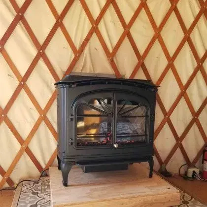 Black wood stove on a wooden platform inside a yurt with tan diamond-patterned walls.