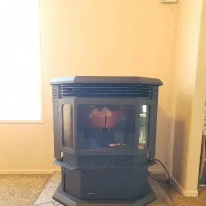 Black wood stove with glass front on a hearth in a beige room near a window.