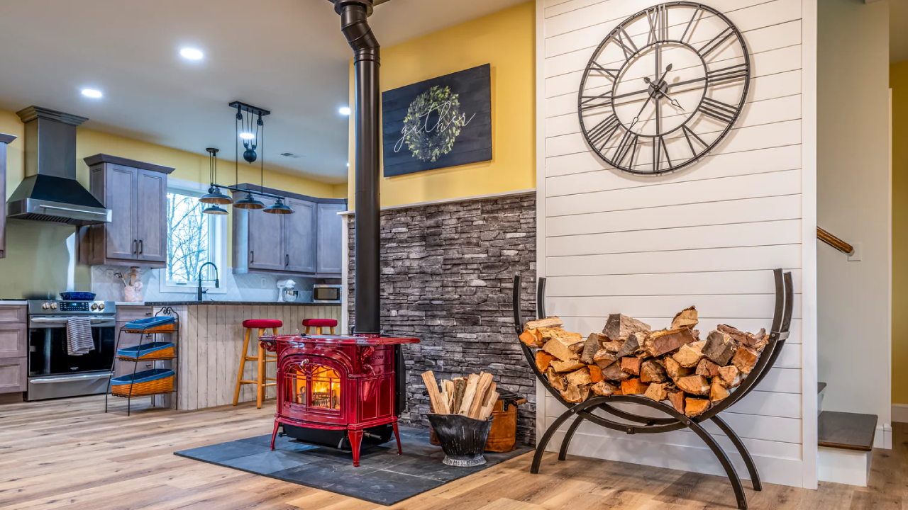 Cozy living room with wood stove, wall clock, red fireplace screen, and kitchen in background