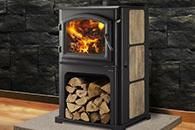 Black wood stove with visible fire and stacked logs inside, set against a stone wall.