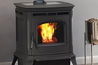 Black cast-iron wood stove with a bright fire burning inside, indoors against a gray wall.