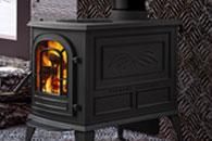 Black cast-iron wood-burning stove with a glowing fire inside, set against a patterned wall.