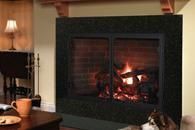 A lit fireplace with glowing logs in a dark green mantel, beside a lamp and side table in a cozy room