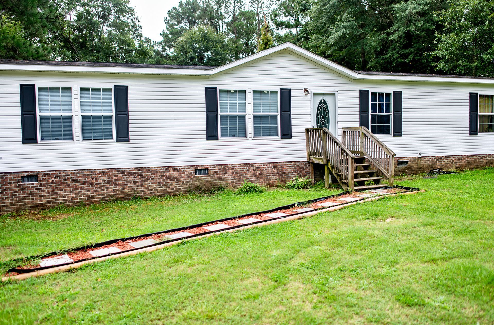 A white mobile home exterior with black shutters, a brick base, and front steps on private property