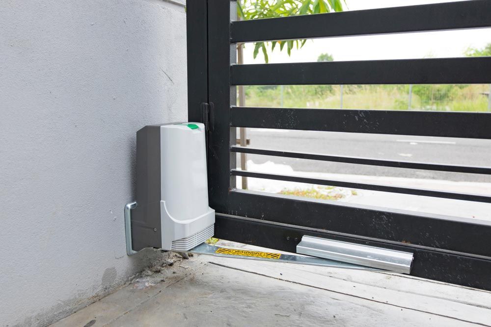 Automatic Gate Opener Attached to a Black Metal Gate and a White Wall — Axcess Garage Doors in Yeppoon, QLD
