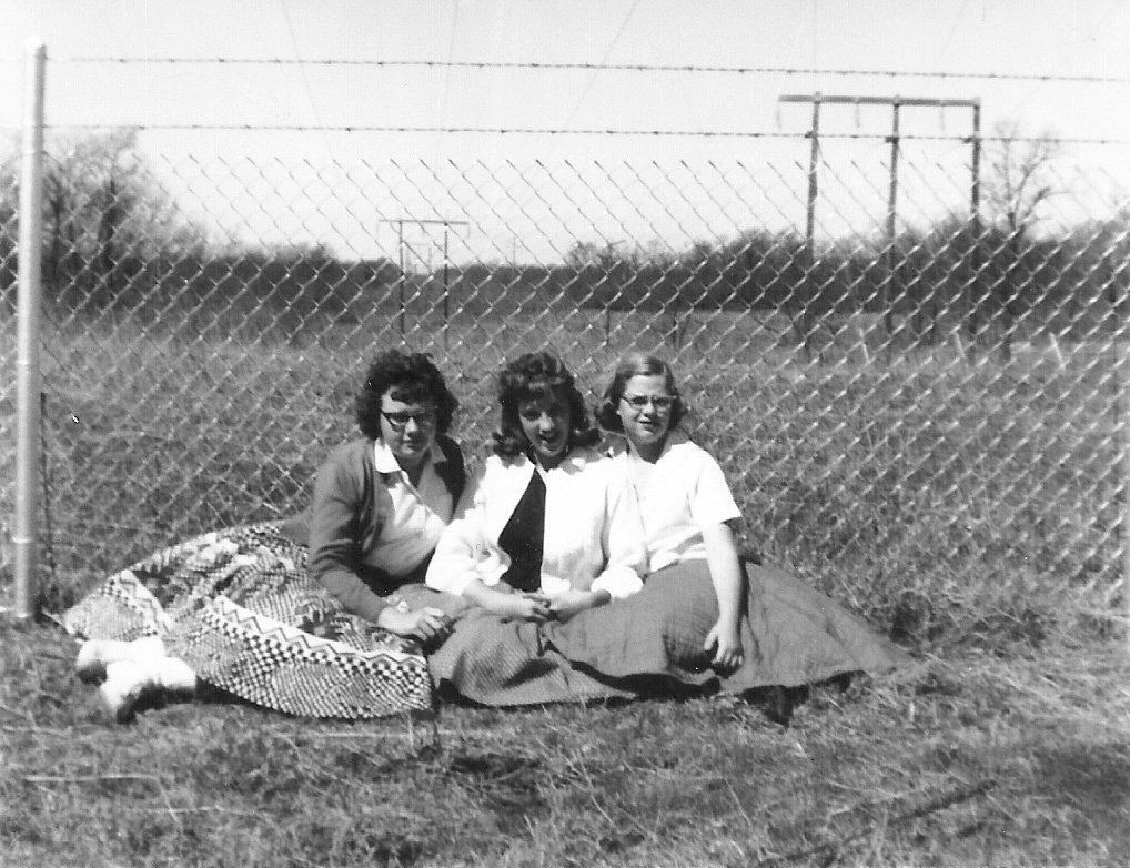 Three women are sitting on the grass in front of a chain link fence.