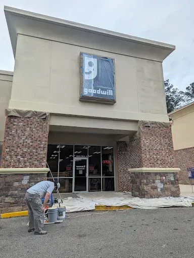 Person painting the entrance of a Goodwill store. Sign is covered in plastic. Exterior brickwork.