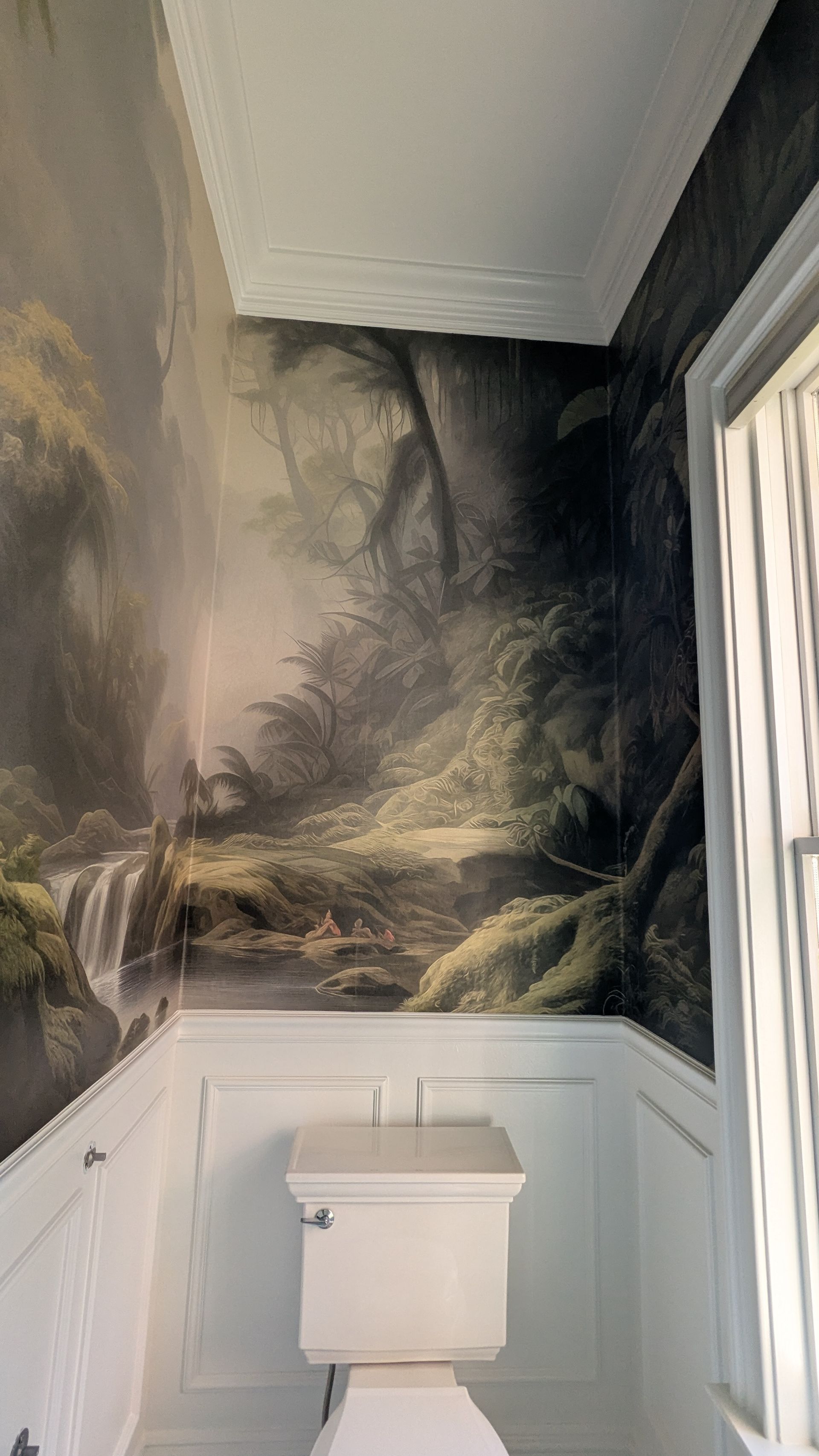 A small bathroom with a forest mural on the wall above a toilet. White wainscoting below.