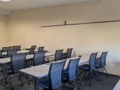 Empty classroom with rows of tables and black chairs. Beige walls, dark carpet.