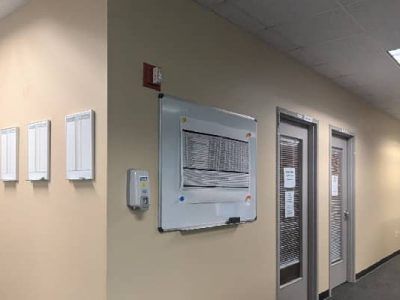 Hallway with a whiteboard, doors, fire alarm, and mounted wall boxes.