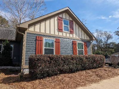 Blue house with red shutters and a brown roof, a hedge in front.