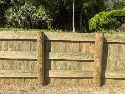 Wooden fence with vertical support posts against a backdrop of green trees and foliage.