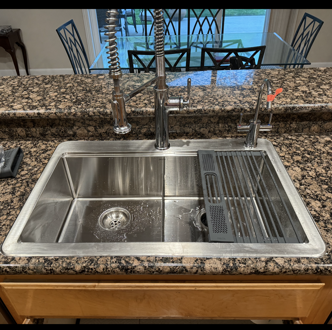 A stainless steel kitchen sink with a spring-coil faucet and a built-in metal drying rack set into a granite countertop.