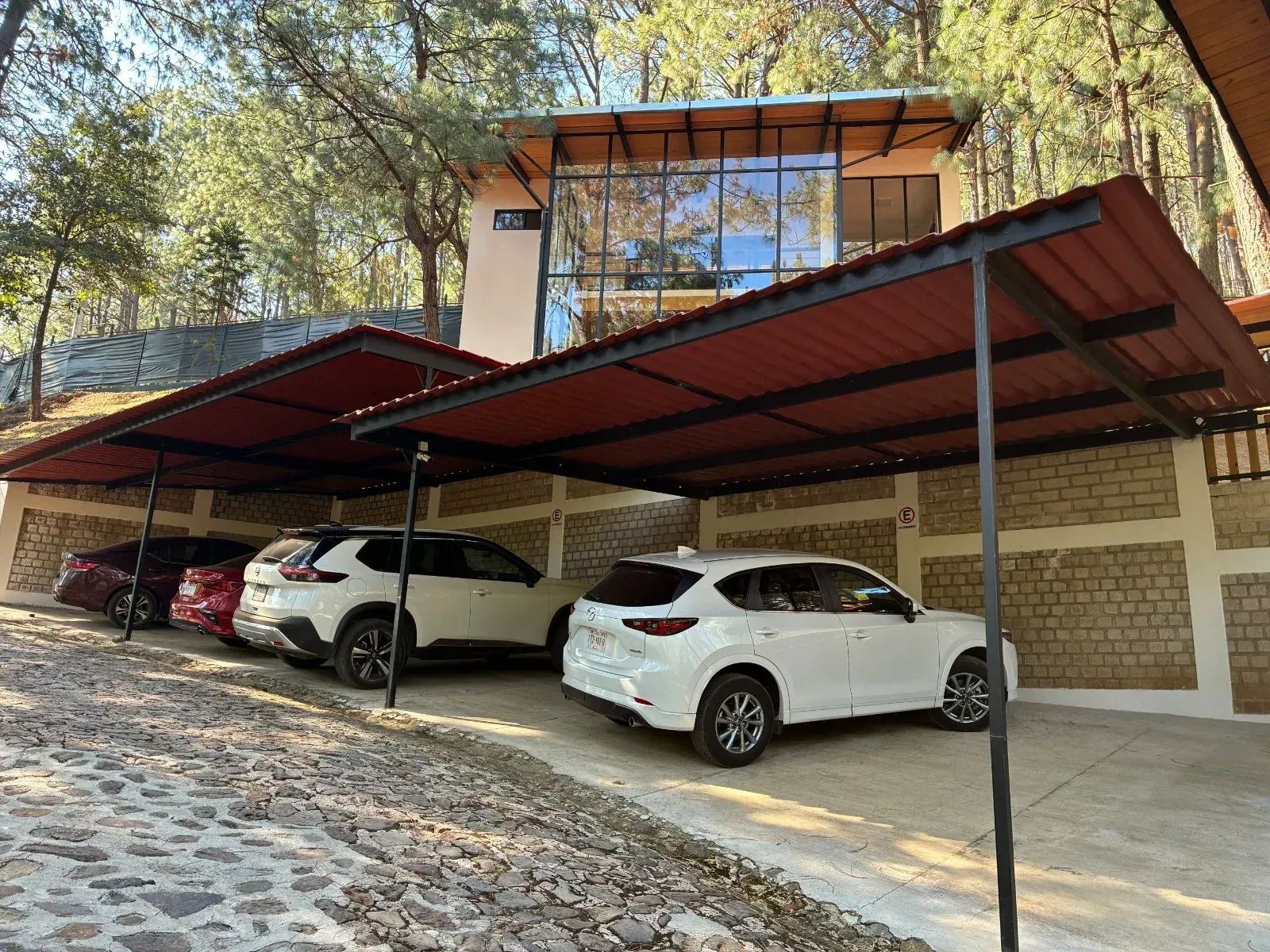Zona de aparcamiento cubierta con tres coches estacionados junto a un edificio de ladrillo y árboles en la azotea.