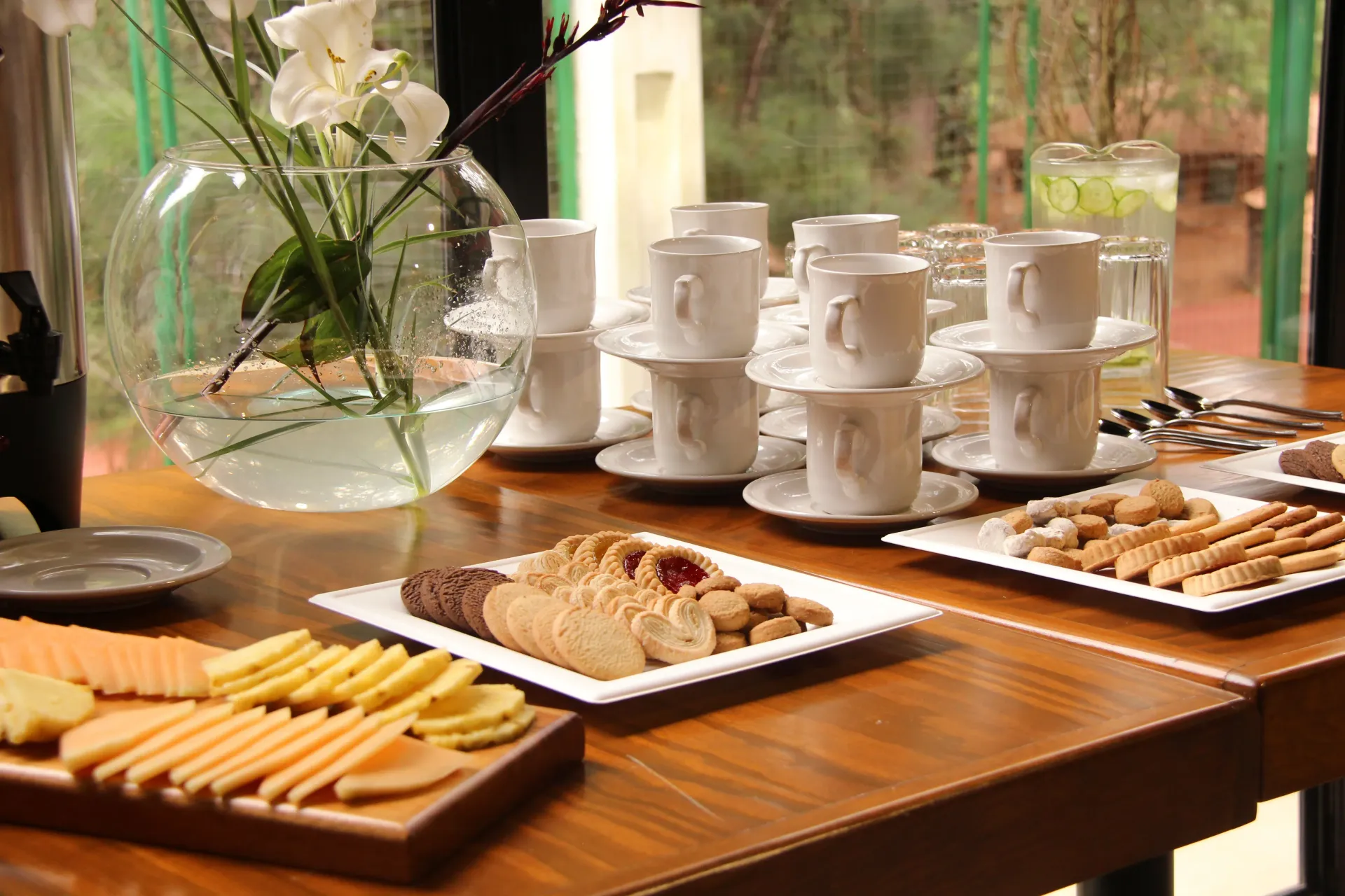Bandejas de pasteles y galletas sobre una mesa de madera junto a tazas blancas apiladas y un jarrón de flores.