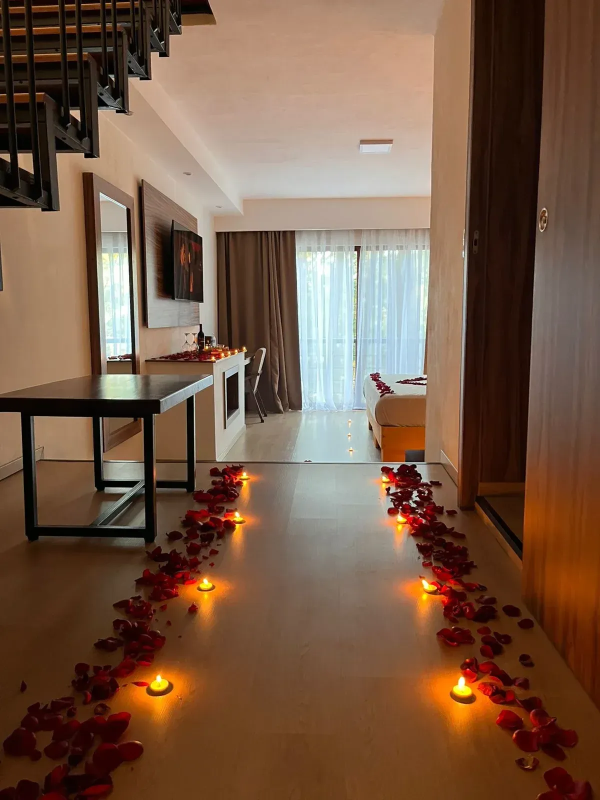 Pasillo decorado con pétalos de rosa y velas encendidas que conduce a un balcón con cortinas transparentes.