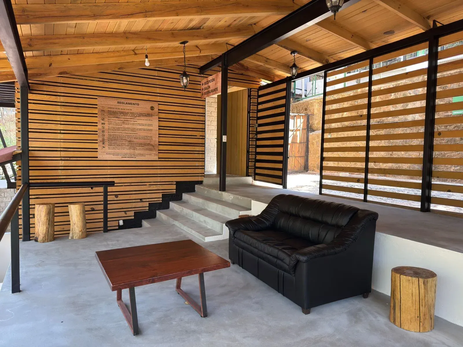 Salón moderno con sofá negro, mesa de centro de madera, paredes de troncos y ventanas a rayas.
