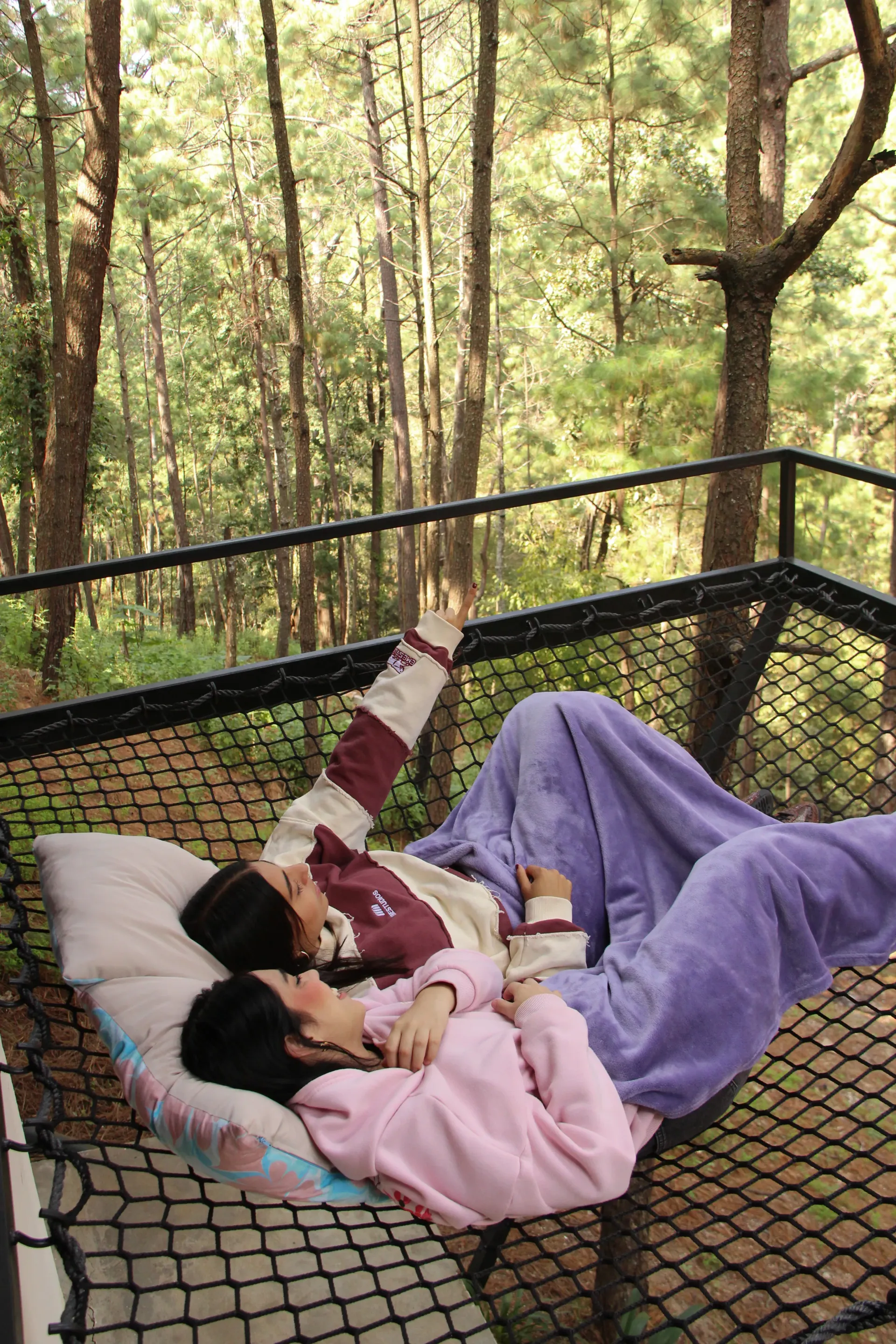 Persona descansando en una hamaca al aire libre, envuelta en una manta morada con un bosque de fondo.
