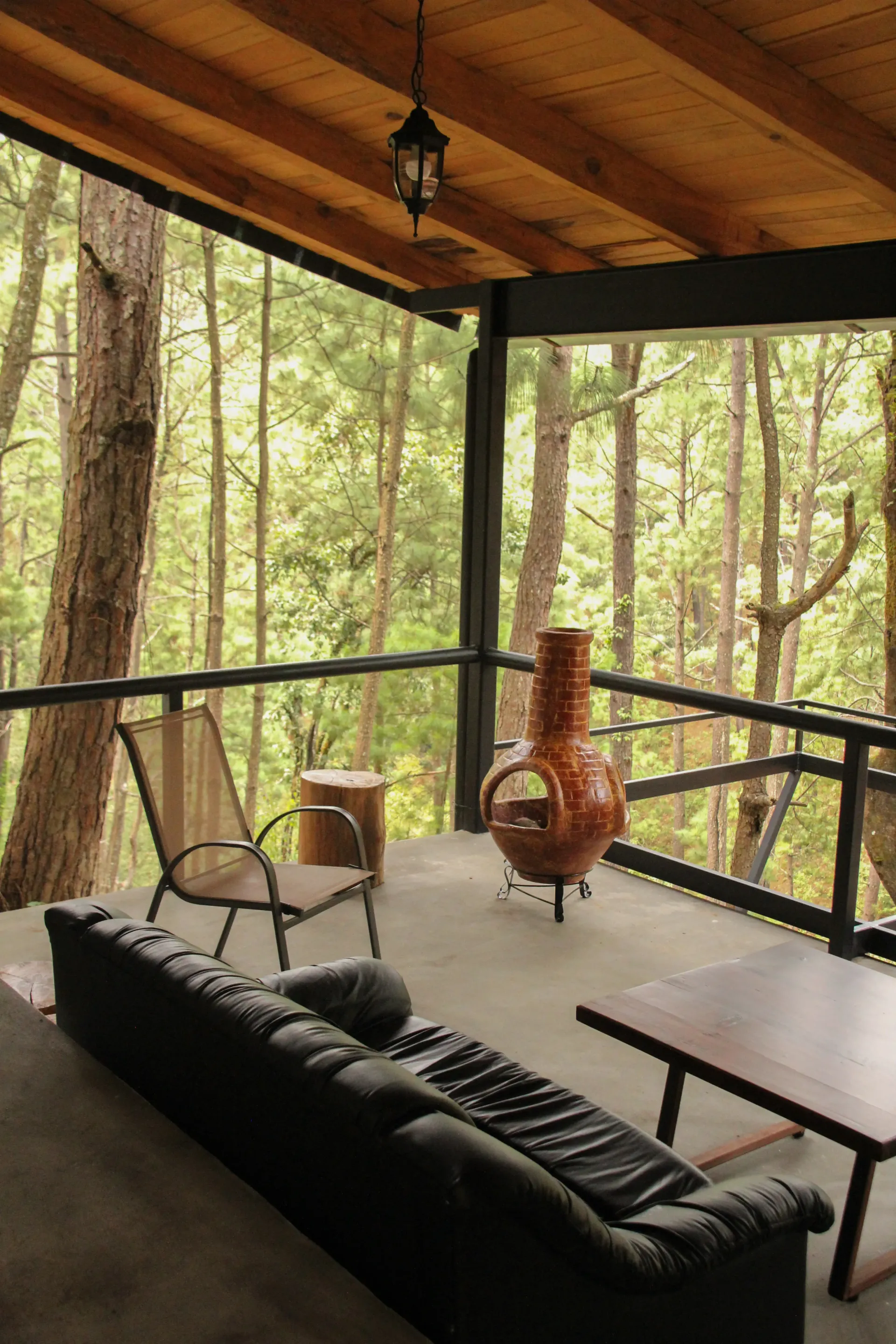 Porche cubierto con sofá negro, silla, mesa y estufa de leña con vistas a un bosque.