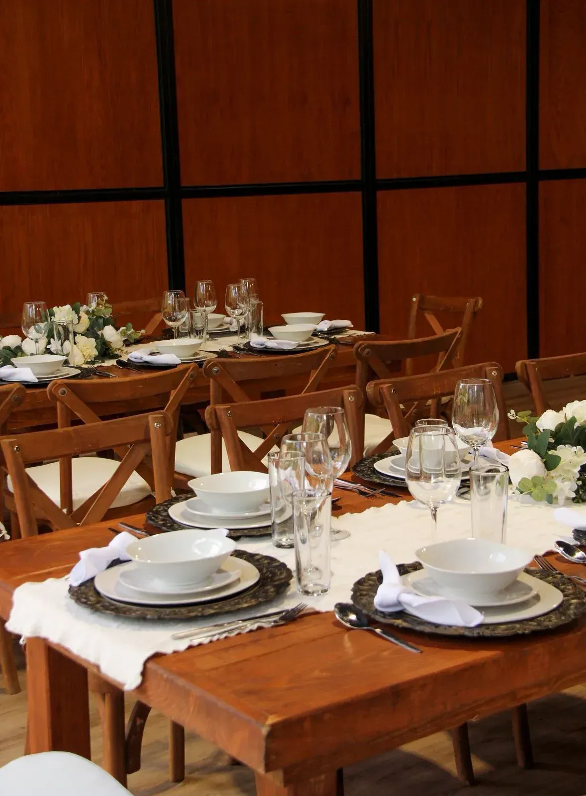 Mesas  para eventos con platos blancos, cristalería y sillas de madera en un salón de banquetes con iluminación cálida.