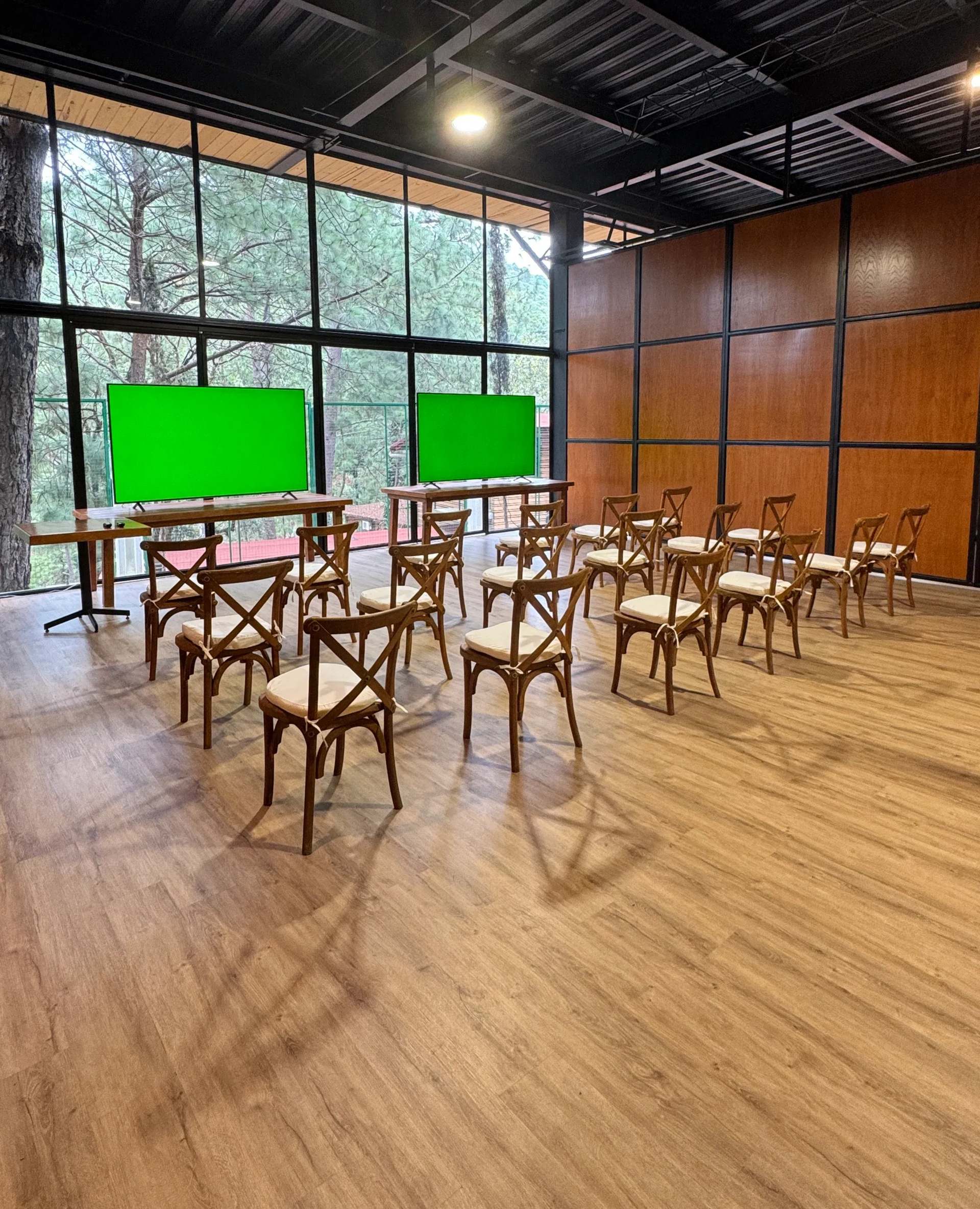 Aula vacía con sillas y pupitres de madera frente a dos pantallas verdes junto a grandes ventanales.