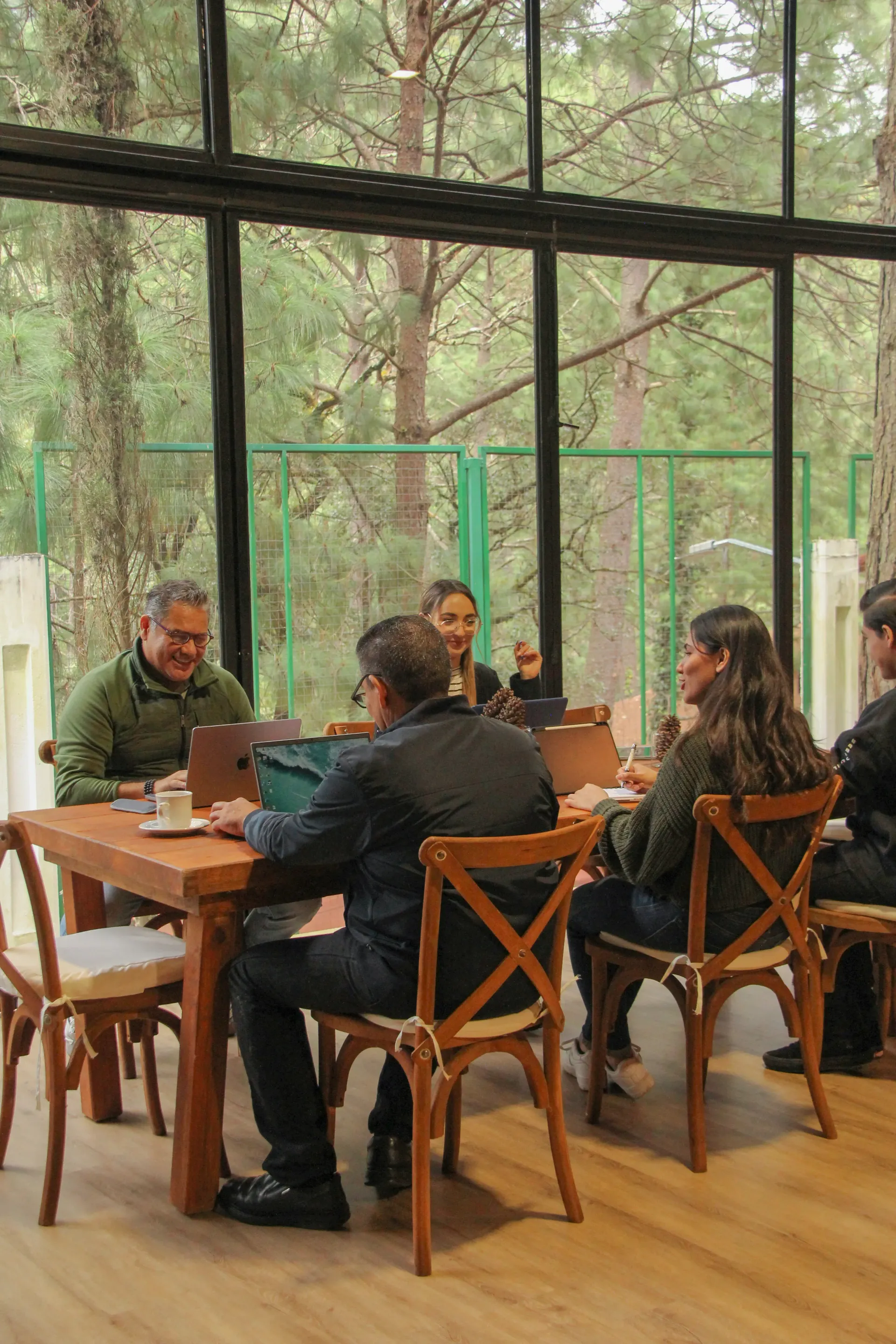 Personas están sentadas alrededor de una mesa de madera en una cafetería con paredes de cristal y árboles en el exterior
