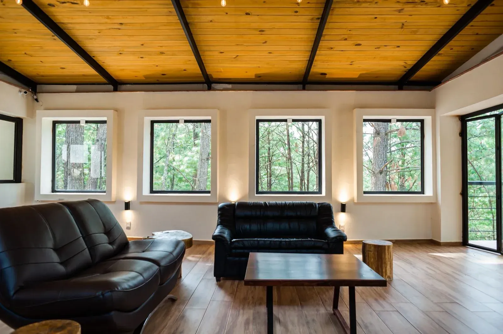 Sala de estar moderna con sofás negros, mesa de centro, grandes ventanales y techo revestido de madera.