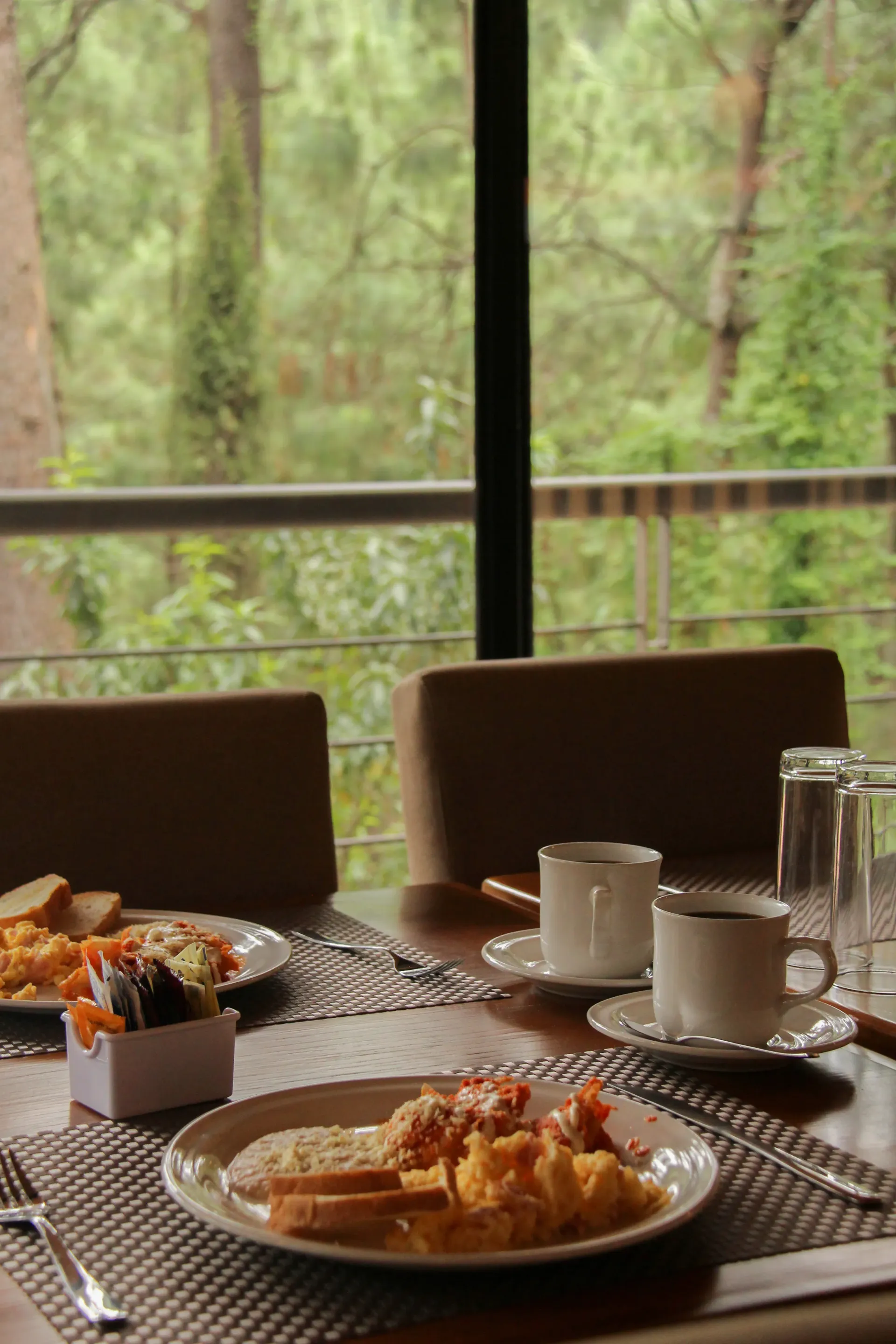 Mesa de desayuno puesta junto a la ventana con platos, tazas de café y vistas al bosque.