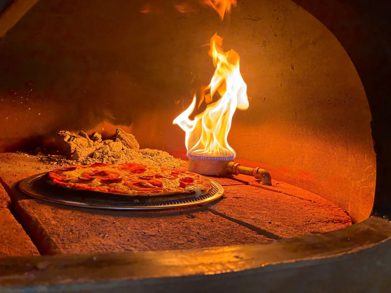 Pizza horneándose en un horno de leña con llamas y una pala para pizza en el interior.
