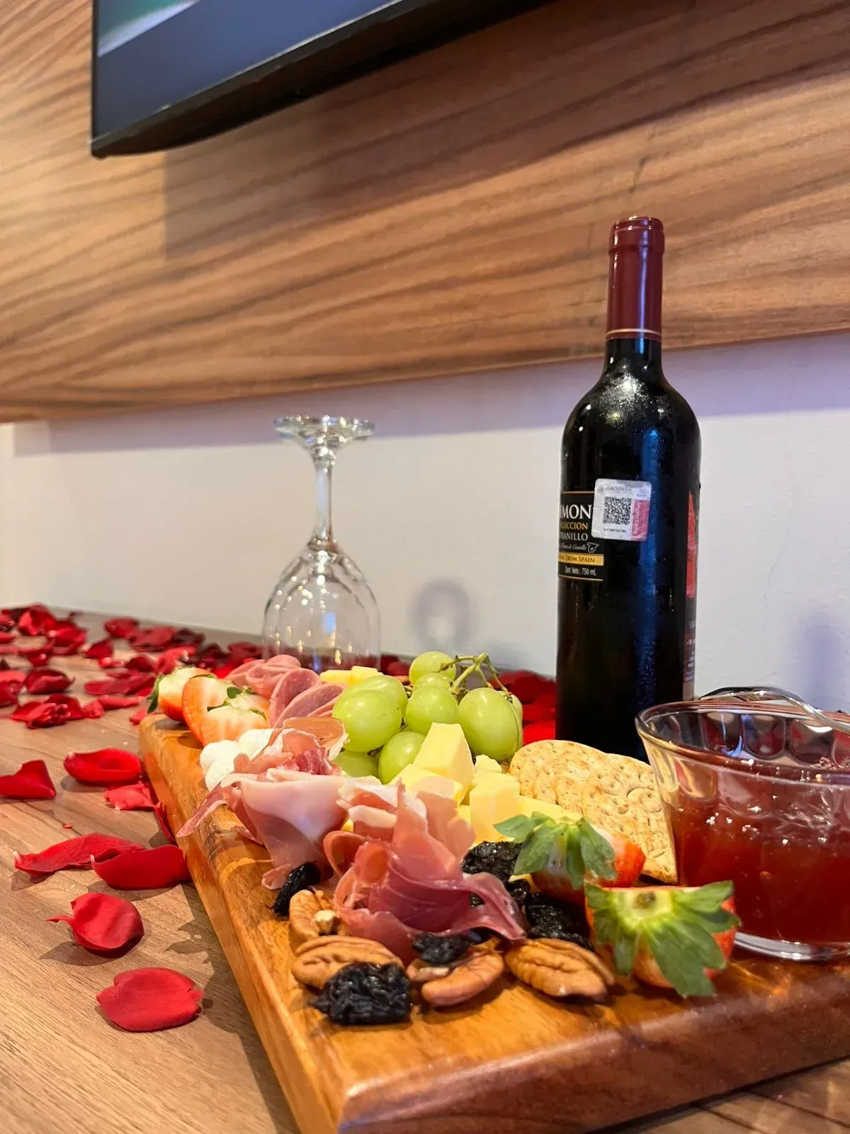 Tabla de embutidos con fruta, queso y galletas saladas junto a vino tinto sobre una mesa con pétalos de rosa.