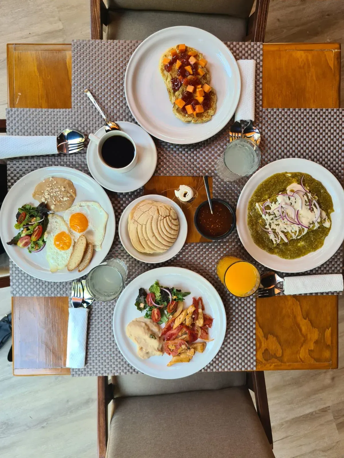 Vista desde arriba de una mesa de desayuno con huevos, café, pasteles y platos servidos sobre manteles individuales tejidos.